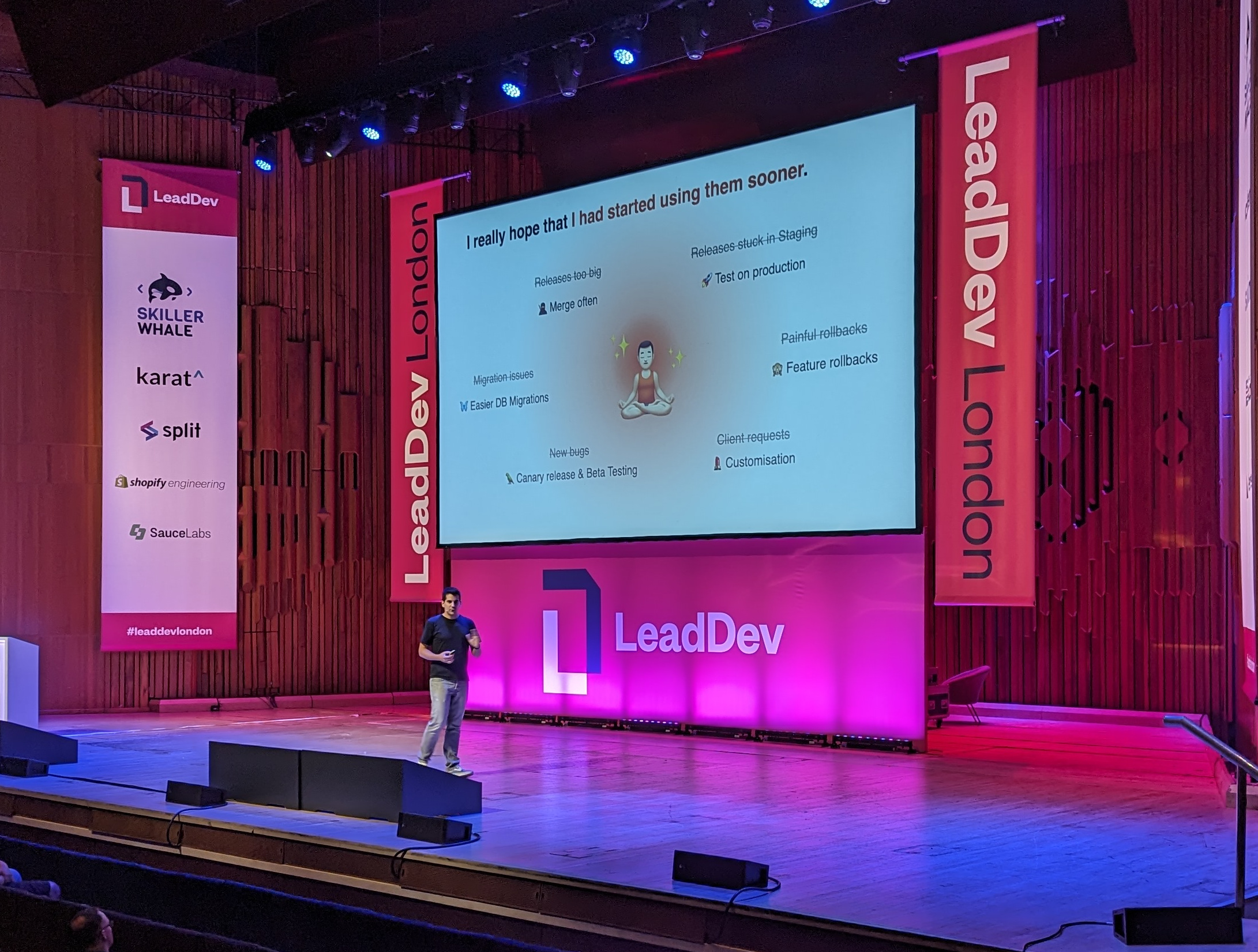 Roger on the stage at LeadDev in front of a slide with a cloud of words around an emoji of a person meditating, with the text "releases too often" crossed out and replaced with "merge often", with the text "migration issues" crossed out and replaced with "easier DB migrations", with the text "new bugs" crossed out and replaced with "canary release and beta testing", with the text "releases stuck in staging" crossed out and replaced with "test on production", with the text "painful rollbacks" crossed out and replaced with "feature rollbacks", and with the text "client requests" crossed out and replaced with "customization"