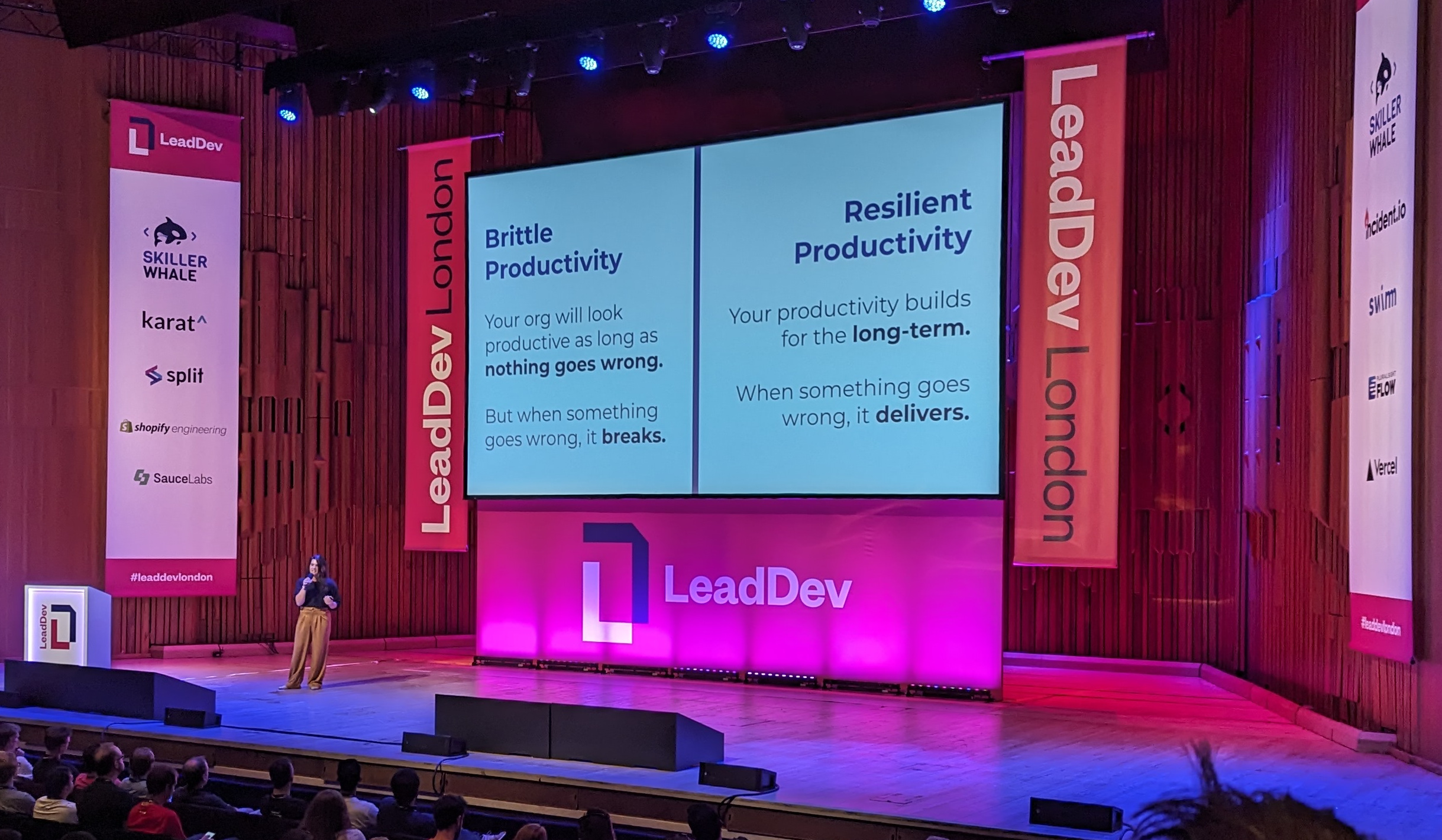 Cat Hicks on the stage at LeadDev in front of a slide that's split in two. On the left hand side, the title "Brittle Productivity", and under it "Your org will look productive as long as nothing goes wrong. But when something goes wrong, it breaks". On the right hand side, the title "Resilient Productivity" and under it "Your productivity builds for the long-term. When something goes wrong, it delivers"