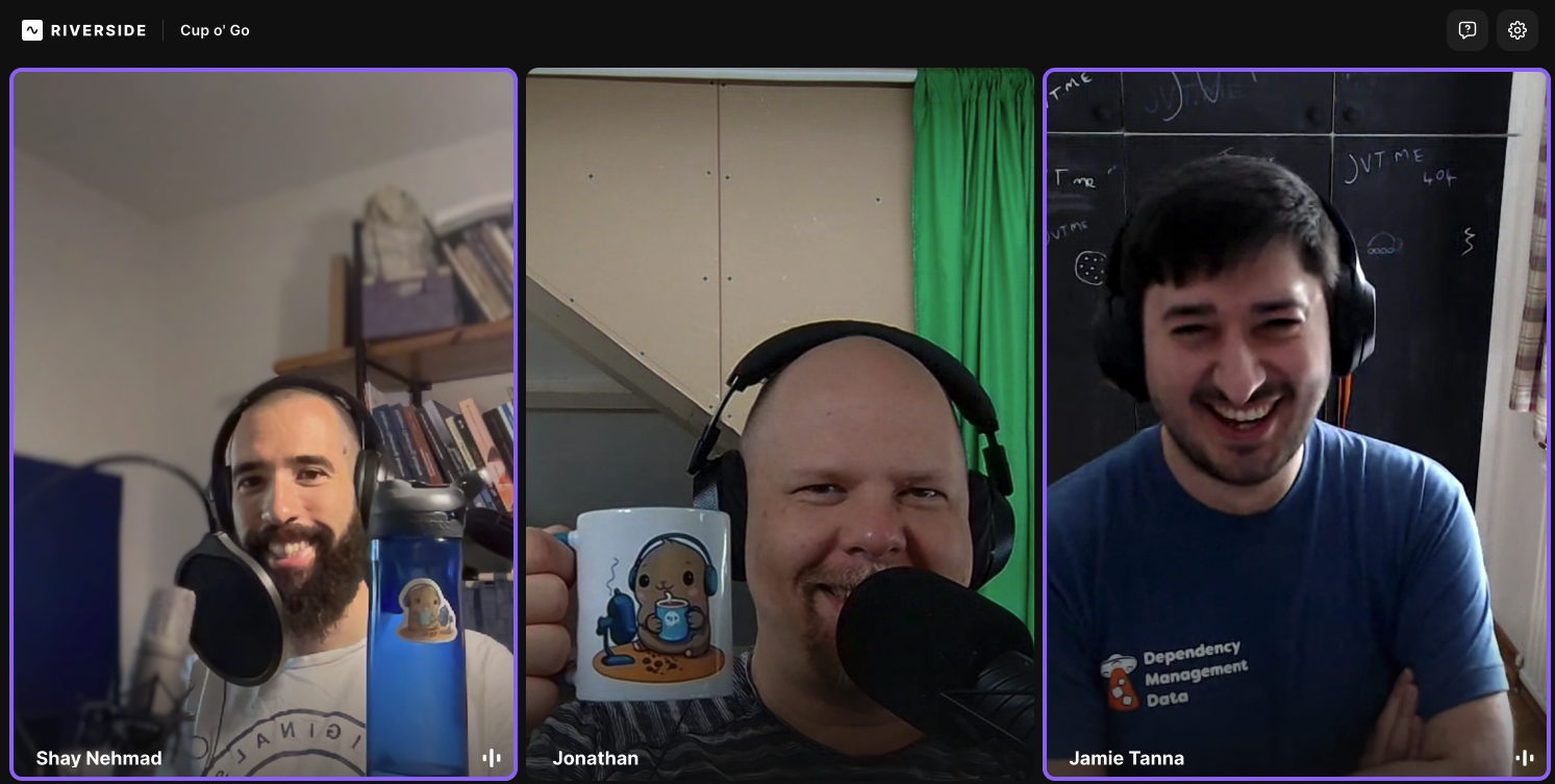A screenshot of the Riverside recording studio showing Shay, Jonathan and Jamie smiling at the camera. Shay is holding up his water bottle, branded Cup o' Go's mascot Brewster, and Jonathan is holding up a Cup o' Go mug. Jamie unfortunately doesn't have any swag (yet) but is wearing his dependency-management-data t-shirt, which he thinks still kinda counts