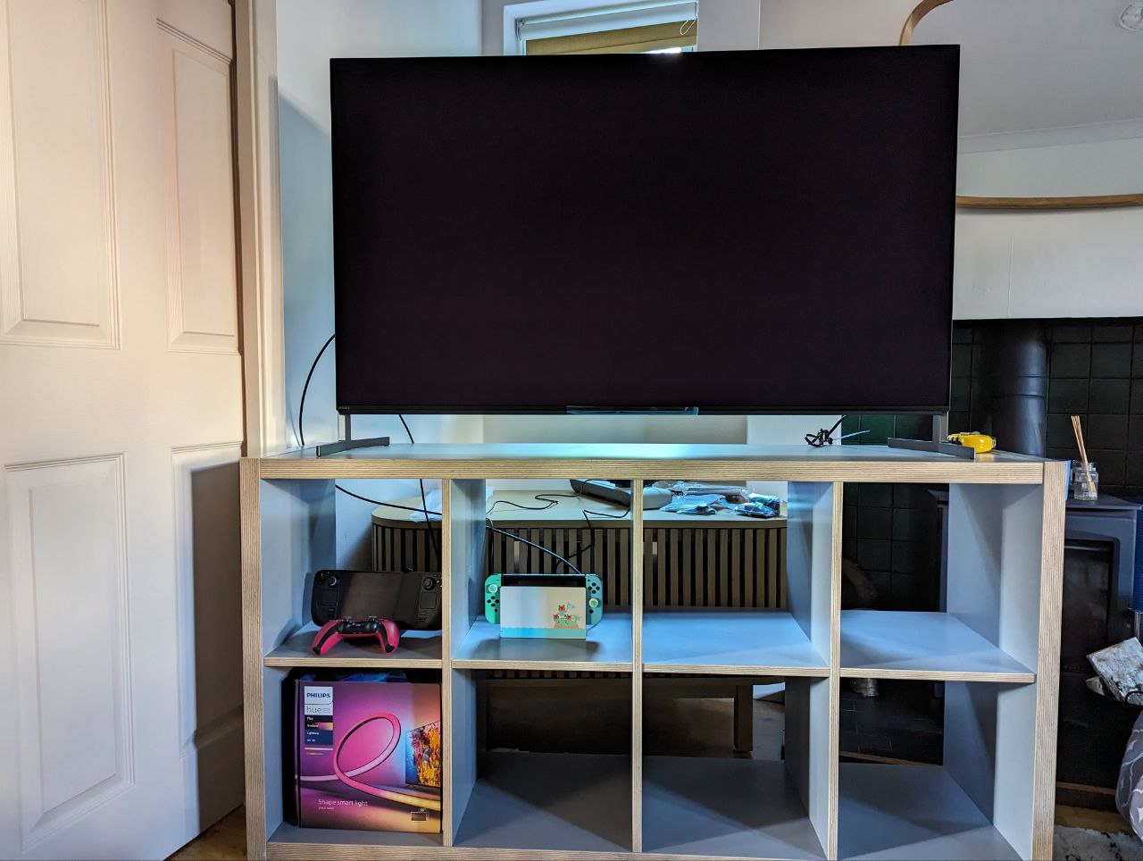 A 55 inch Sony Bravia TV on top of an IKEA Kallax shelving unit, which is laid horizontally a little awkwardly. Behind the Kallax, you can see the new TV stand which is not wide enough to hold the TV, which annoyingly has legs that are the width of the TV, instead of being in the centre