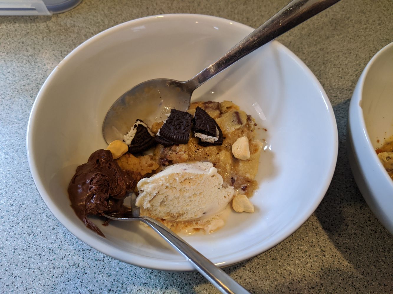 A bowl with half a cookie, a small scoop of ice cream, chunks of honeycomb and some broken up Oreo, and a spoon of Nutella