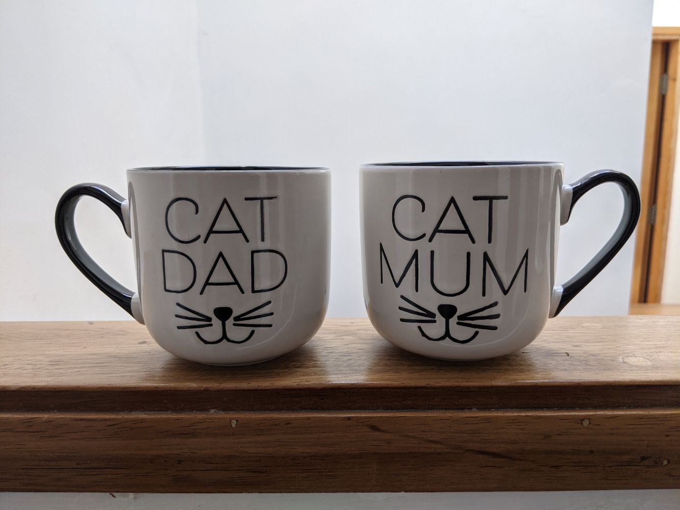 Two mugs sitting on the side, one with "cat dad" and one with "cat mum" and each with a smiling cat mouth 