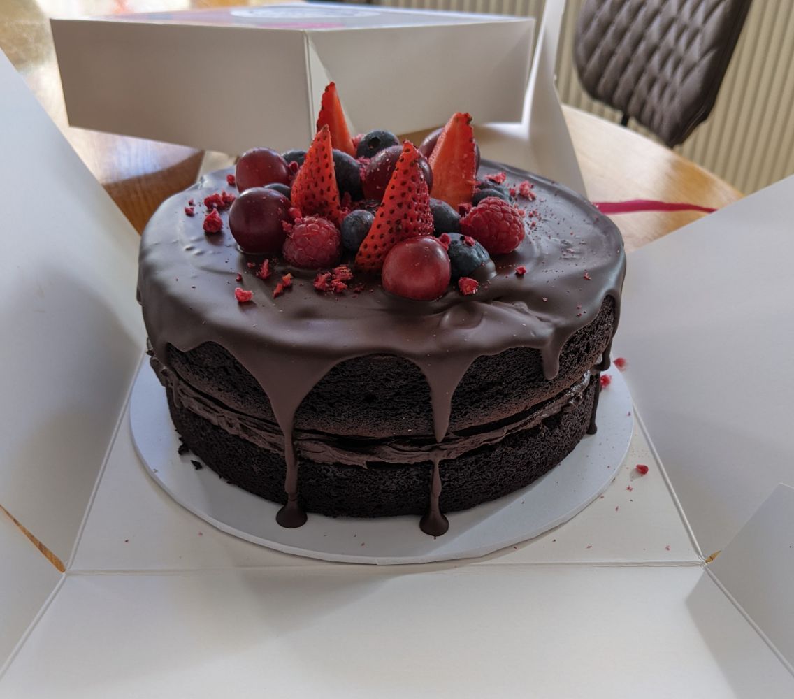 The container of cake opened up, showing a very high chocolate cake with an assortment of strawberries, grapes and raspberries on top, with drips of chocolate icing down the side of the cake