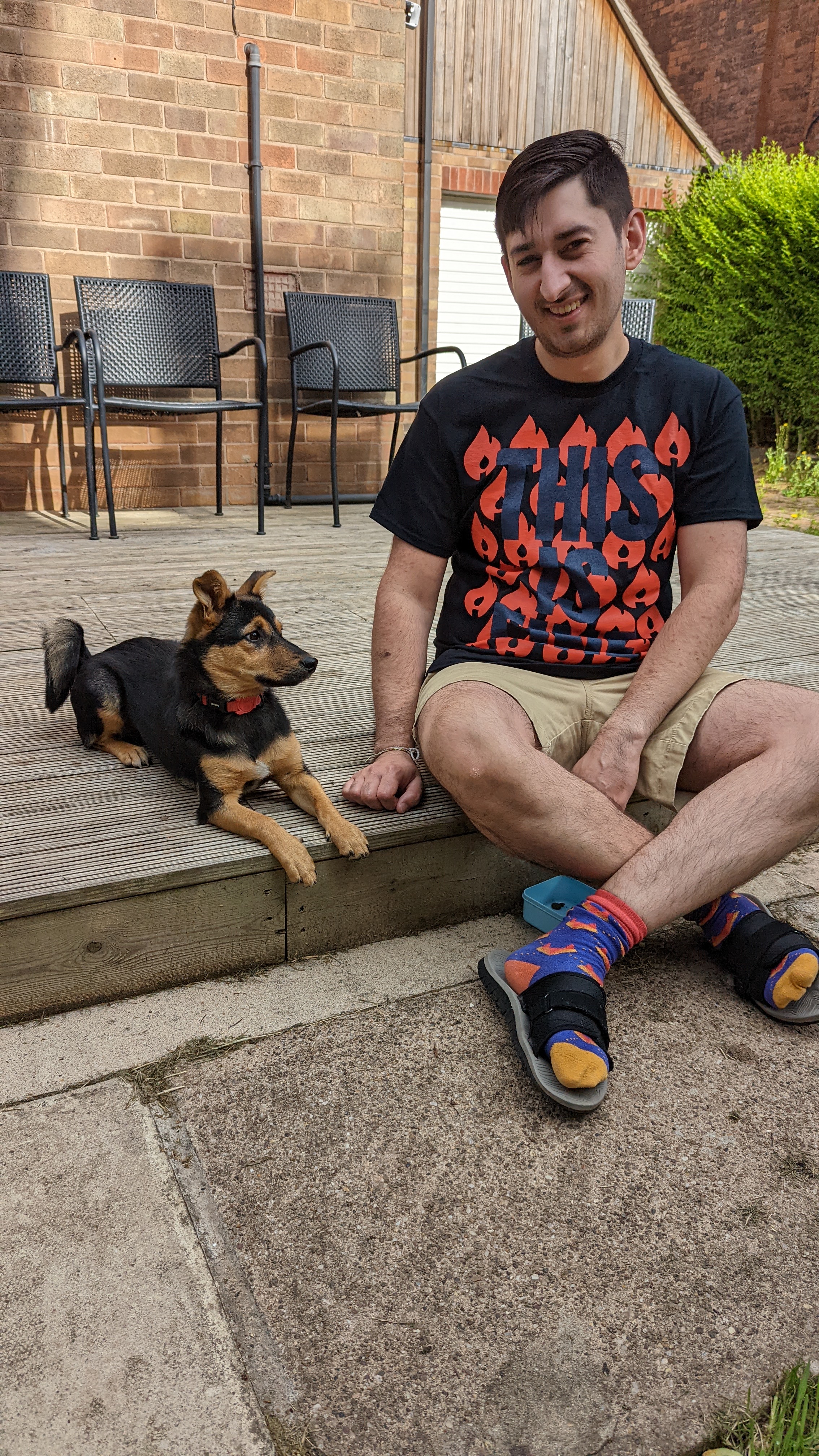 Jamie and Cookie sitting outside on the decking, with Cookie on Jamie's left and looking away from the camera. Jamie is smiling at the camera and wearing an incident.io t-shirt with lots of fires (their logo) and the words 'this is fine' over them