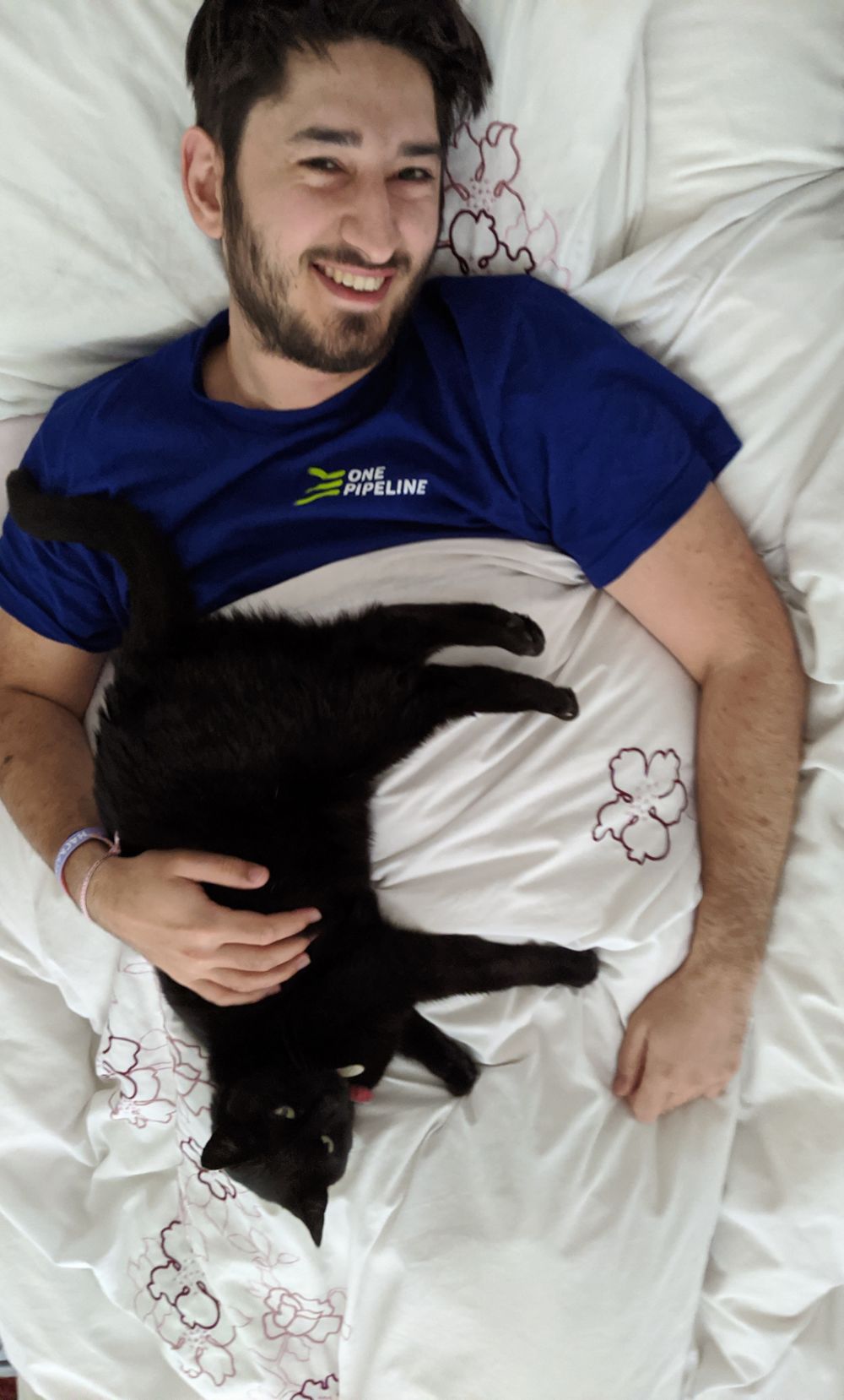 Photo from above of Jamie lying on bed, smiling, with a black cat lying on his belly