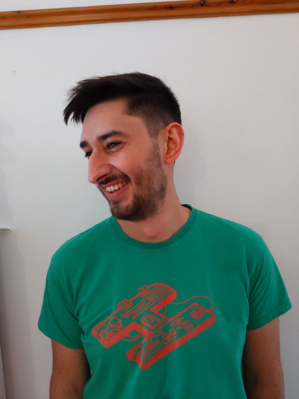 Jamie smiling and slightly looking away from the camera so you can see his new haircut more appropriately - a short haircut combed over with an undercut, and a trimmed beard - while wearing a Hack the Holidays hackathon t-shirt