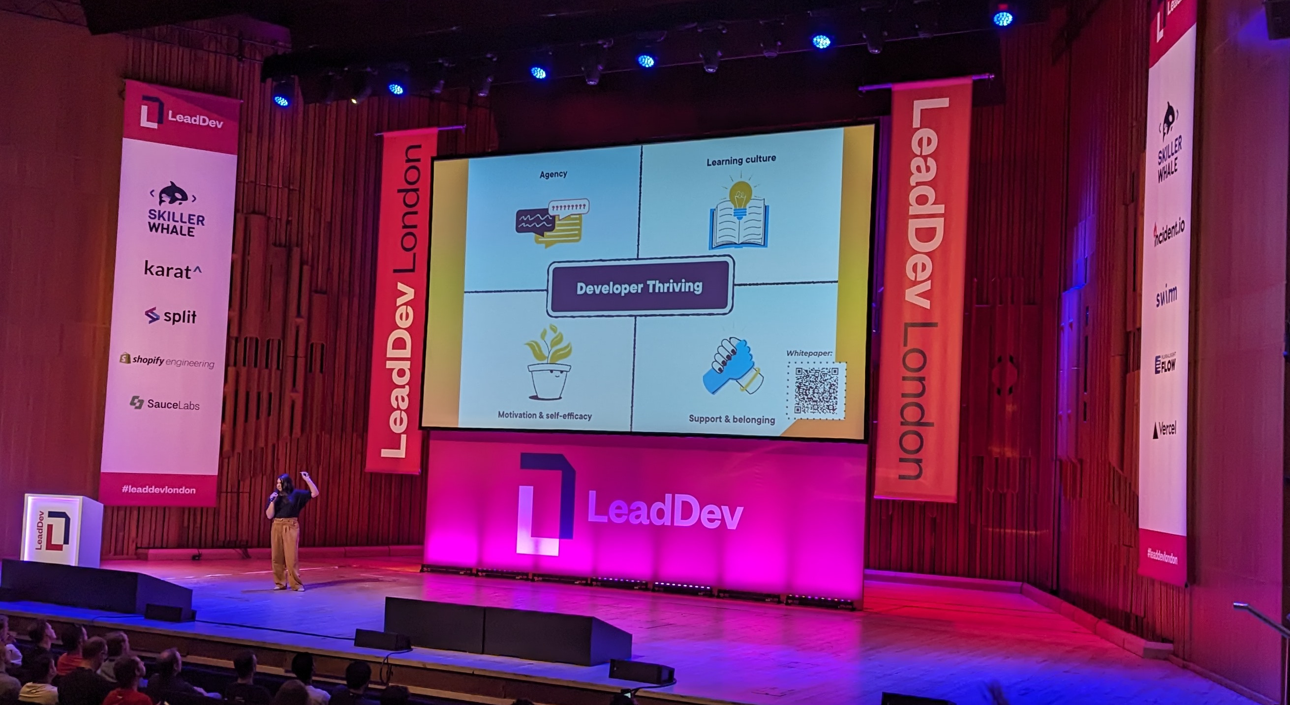 Cat Hicks on the stage at LeadDev in front of a slide split up into four quadrants around the title "Developer Thriving", with each quadrant having an illustration that aligns with the title. The quadrants are Agency, Learning culture, Motivation & self-efficacy and Support & belonging