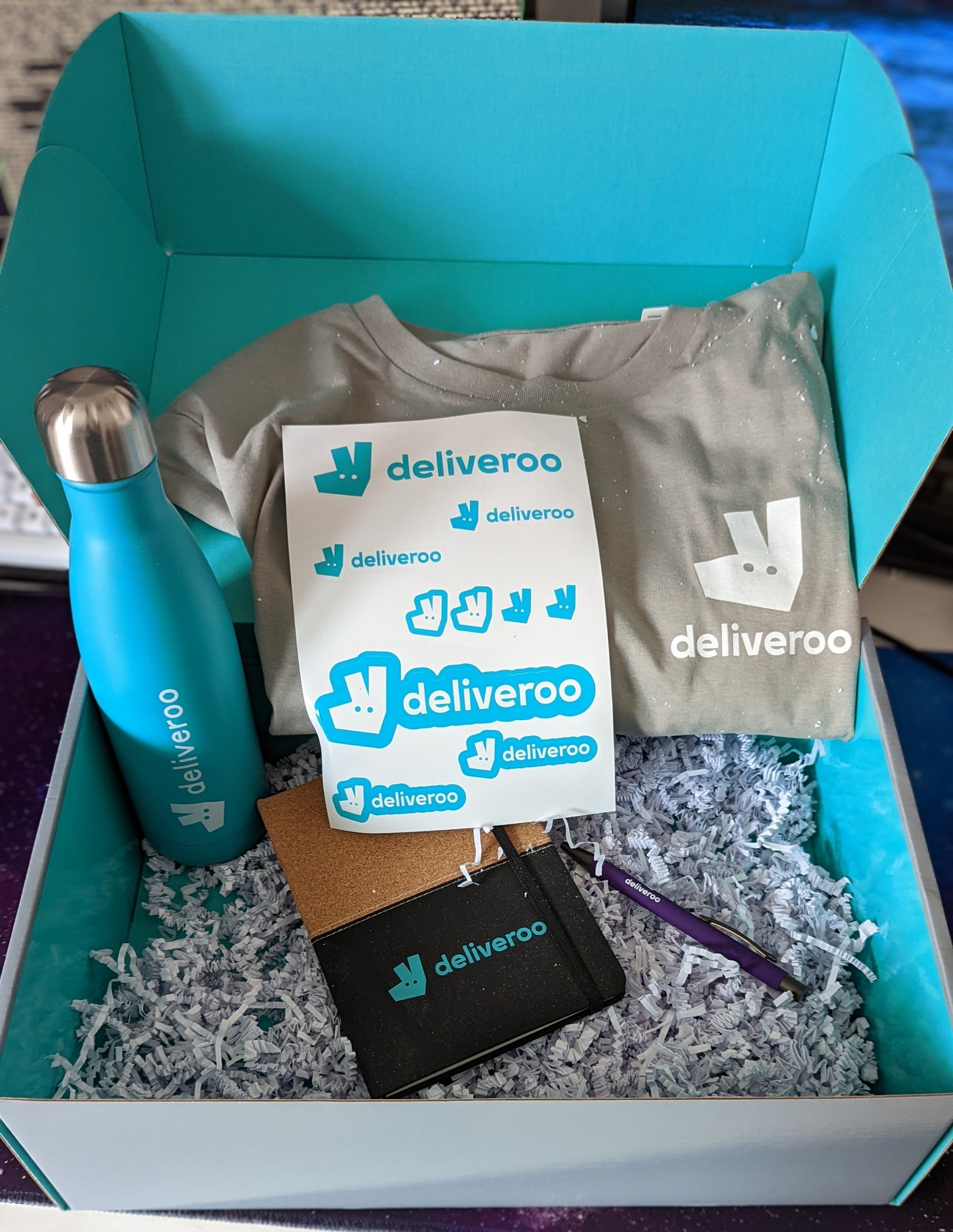 The inside of the box with all the swag visible - a water bottle in Deliveroo blue, a set of (laptop) stickers of the logo and name, a Cork and black notebook, a pen, and a Deliveroo brown t-shirt