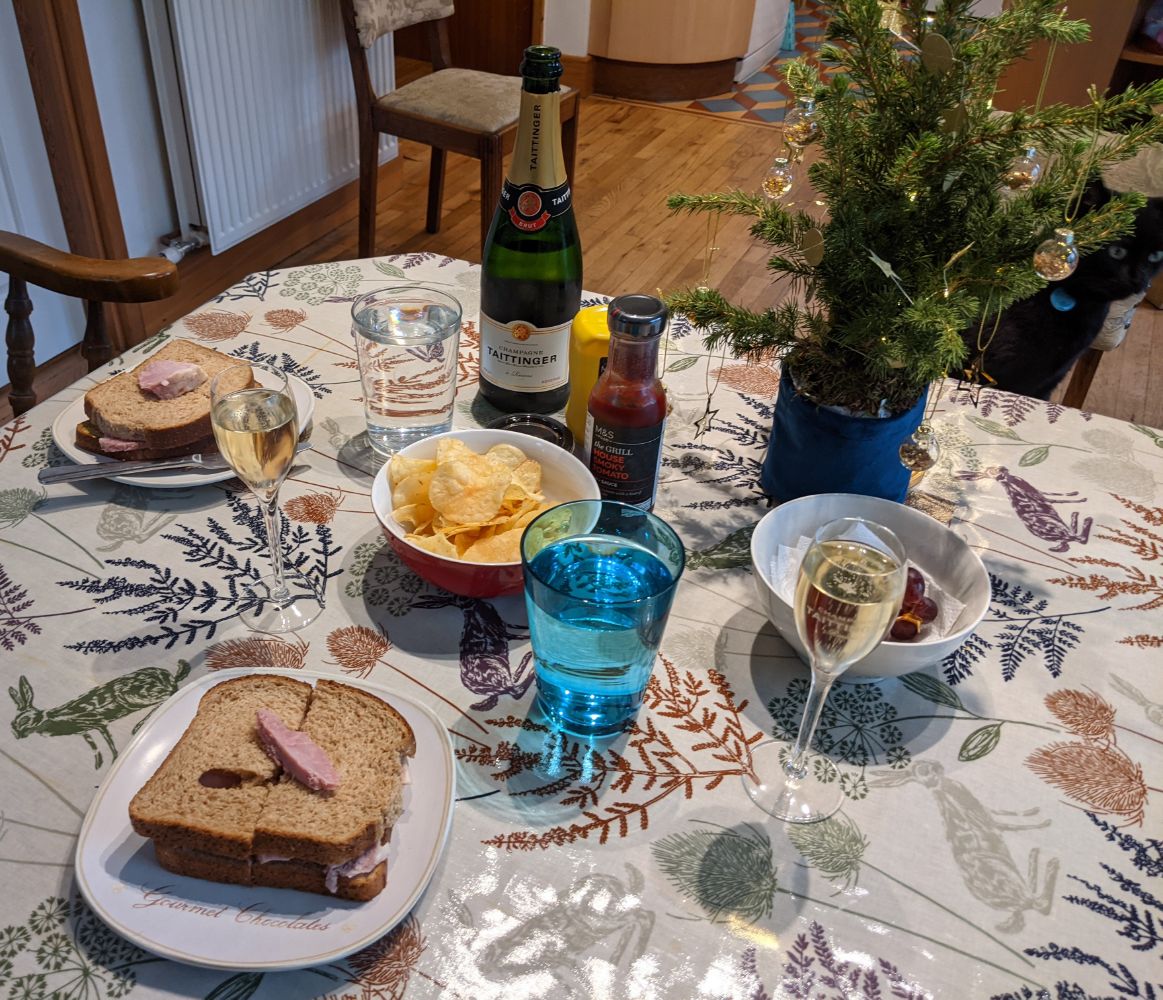 A table laden with food, with two sandwiches filled with marmalade glazed ham and M&S' fancy ketchup, with a slice of ham on top for good measure, Wensleydale cheese and cranberry crisps in a bowl, two glasses of Tatinger Champagne, a small Christmas tree from Bloom and Wild, a bowl of red grapes, and a black cat in the background looking away from the camera