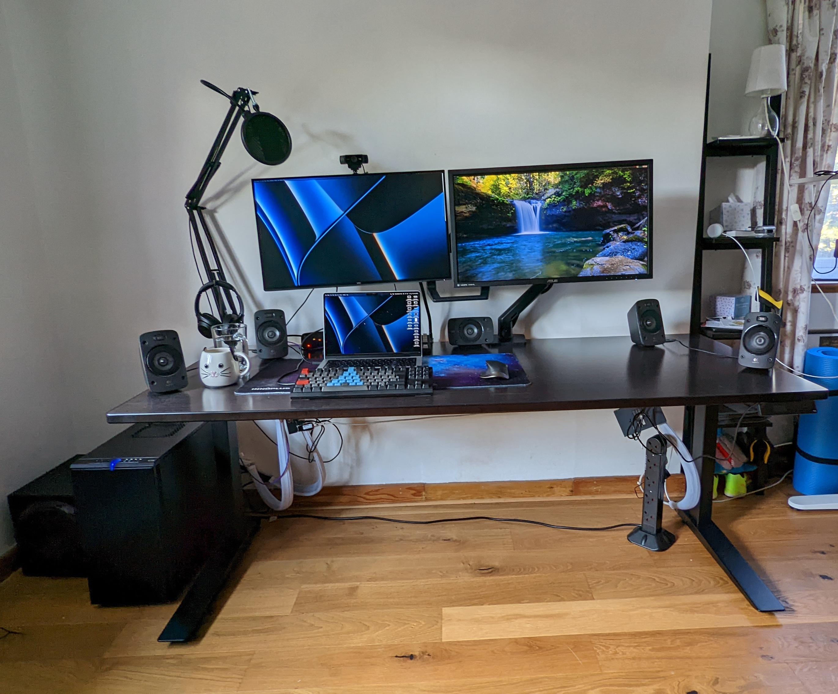 A photo of a Jamie's desk setup in "work" mode. The desk is a Fully Jarvis sit-stand desk, with a dark bamboo top and black legs. Around the desk position is a set of surround sound speakers. There is a microphone arm in the left hand side corner of the desk, and a headphones stand in front of it. On the desk, there is a nebula themed large mouse-and-keyboard pad, on top of which is a WASD Keyboard v2 keyboard (in dark grey, black, red and light blue colours, where the blue keys are in the shape of the Arch Linux logo), a Logtech MX Master mouse, and a Deliveroo-issued M1 Macbook Pro. There are two screens, using an Ergotron dual monitor mount. The left hand side screen is a Dell UltraSharp (4k) 27" USB-C Hub Monitor - U2722DE, and the right hand side screen is an Asus PB287Q 28" 4K Monitor. Above the left hand side monitor is a Logitech C922 webcam. On the left hand side screen, the background image is the same as the Macbook, indicating that it is connected to the laptop, and on the right, the image is a waterfall into a natural enclosed pool. Below the desk you can see an Ethernet switch on the right which unfortunately isn't yet cable tidied, as well as a USB hub that's also visible and not-yet-tidied, on the left. There is also a little bit of mess and some shelves on the right hand side of the image that couldn't be cropped out easily.