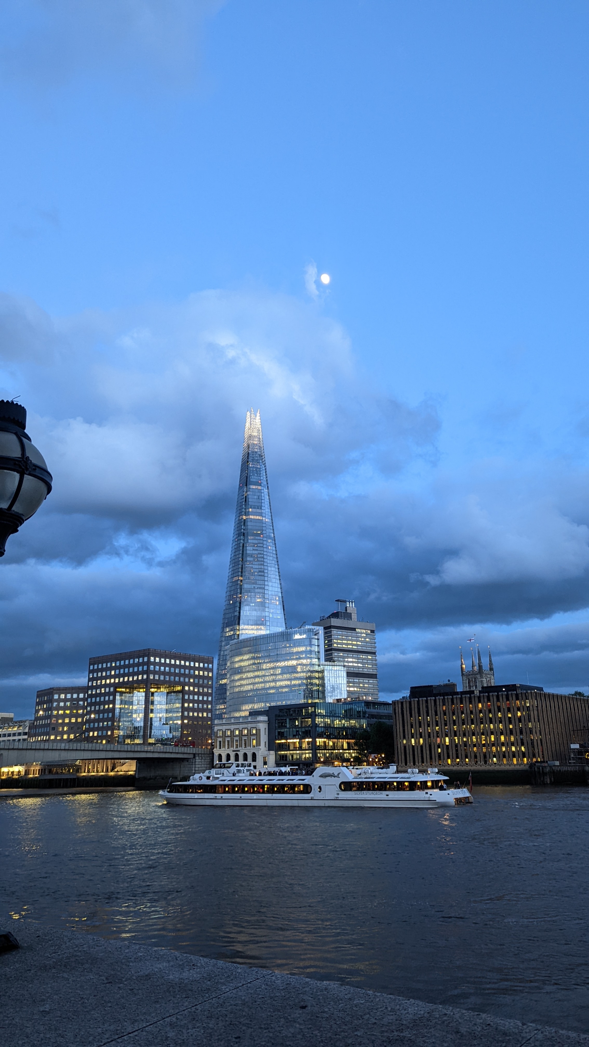 A late evening view of The Shard with clouds above it and just peeking out is the moon, brightly shining. It's a lovely bright photo despite being shot so late in the evening, and a sign of the lovely summer evenings