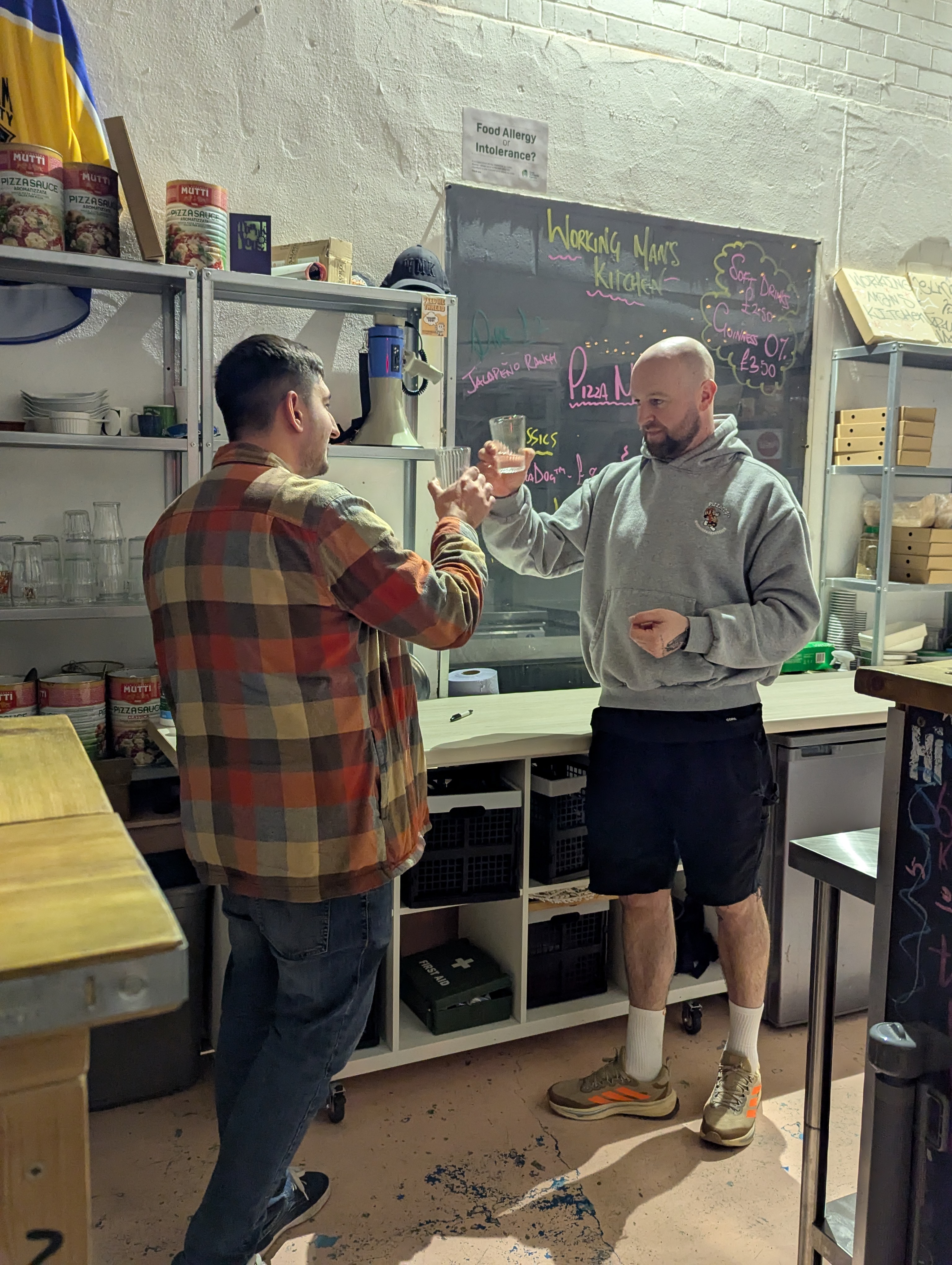 Jamie and Paddy "cheers"ing their glasses of sake. The sake is unfortunately blocked by Jamie, facing away from the camera, who is wearing a very nice and warm overshirt on top of a t-shirt that's not pictured but is really quite nice and you should be sad you can't see it, and Paddy is looking at Jamie, friendly as always