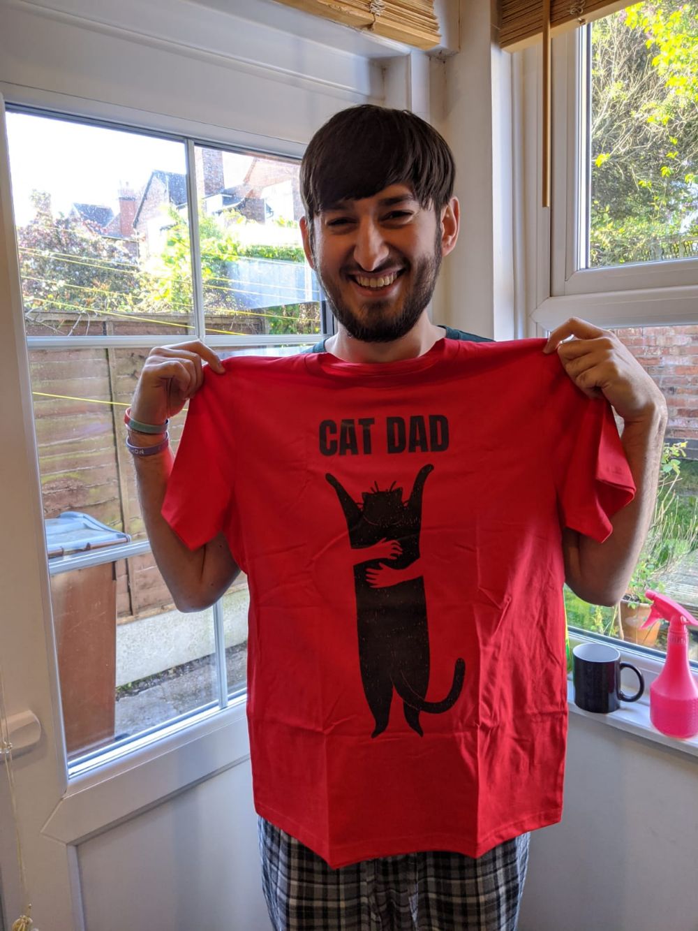 Jamie holding up a red t-shirt with a black cat being cuddled (seemingly against its will) with a title "cat dad"