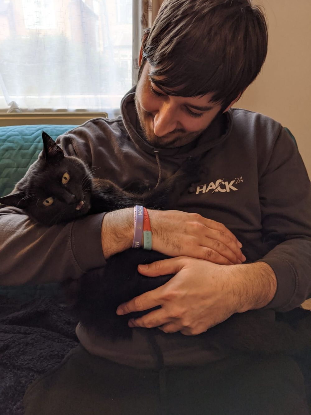 Jamie holding black cat in the crook of his arm like a baby, smiling at him, while the cat meows a little at the camera Jamie holding black cat in the crook of his arm like a baby, smiling at him, while the cat meows a little at the camera