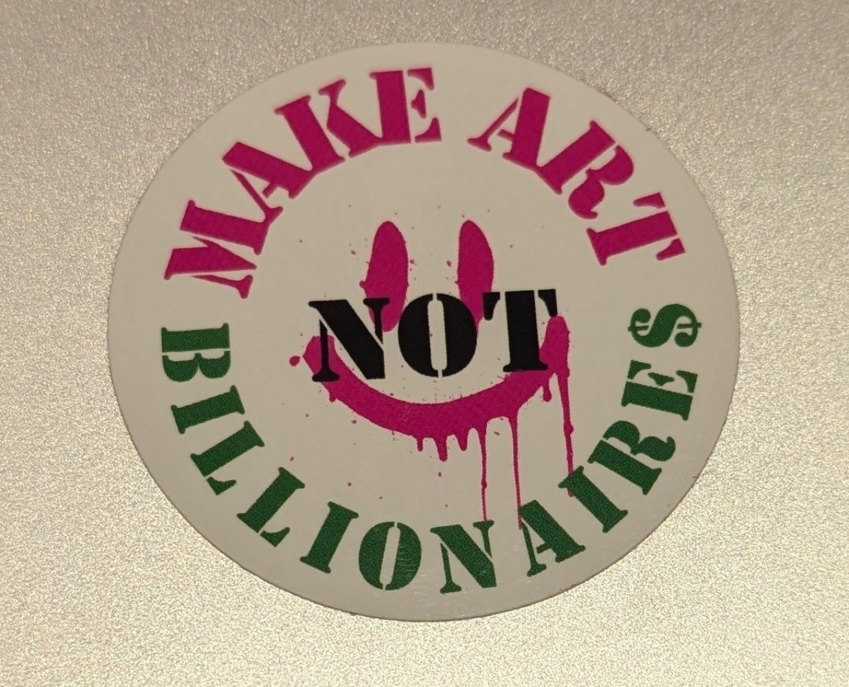 A laptop sticker, placed on Jamie's laptop, saying "make art, not billionaires" with a smiley face under the "not" 