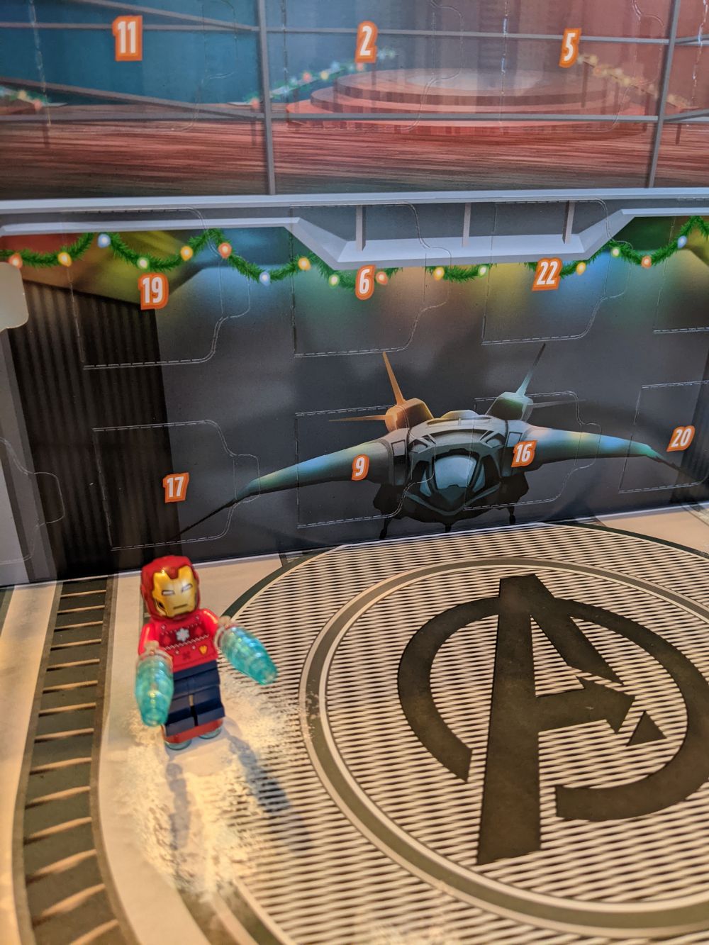 A backdrop of the Avengers hangar with a Quinn Jet in the background, and Iron Man in the front, taken from the first day of the advent calendar. Iron Man is wearing his helmet, has a Christmas jumper with the arc reactor on, is slightly floating in mid-air and has his hand weapons primed