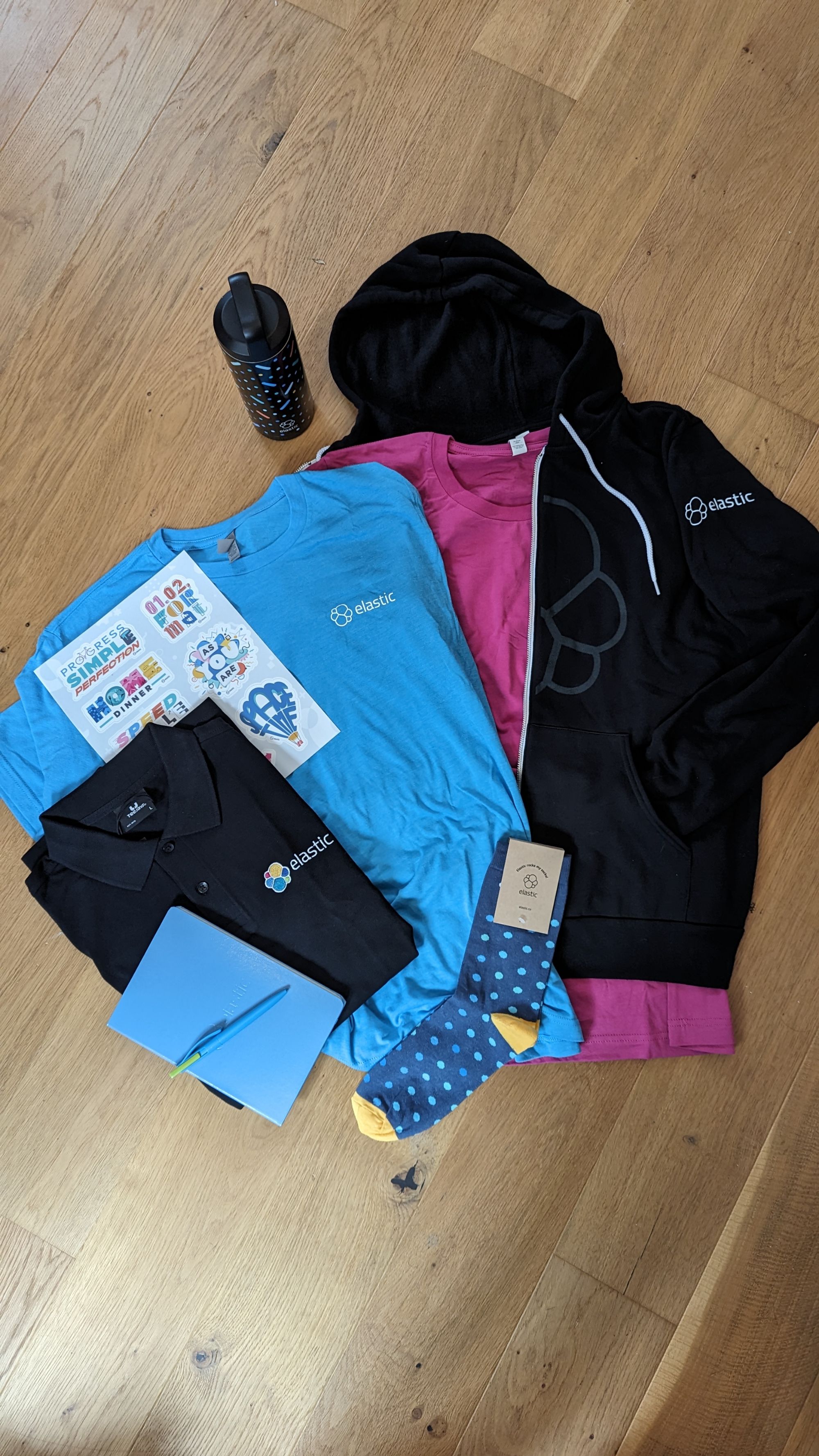 An arrangement of swag from Elastic, arranged neatly, and laid out on the wooden floor. There is a blue notebook, embossed with the Elastic logo, a black polo shirt, a set of stickers with the Elastic values that are super cute and really nicely stylised, a bright blue Elastic, a bright pink Elastic tshirt, a black hoodie, a pair of socks, and a slightly stripy water bottle. All the clothes are incredibly soft, and very high quality. There is also (not pictured) a darker blue "helper node" tshirt for volunteering in