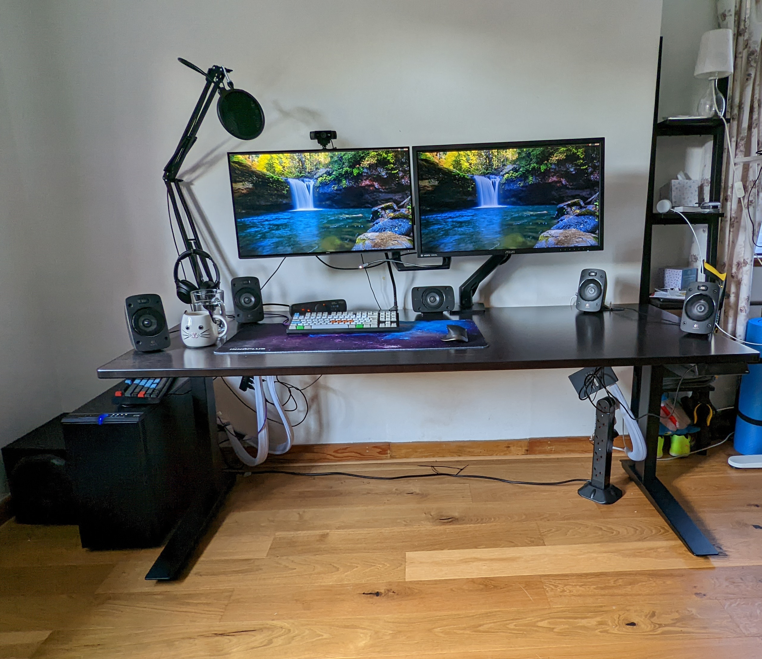A photo of a Jamie's desk setup in "home" mode. The desk is a Fully Jarvis sit-stand desk, with a dark bamboo top and black legs. Around the desk position is a set of surround sound speakers. There is a microphone arm in the left hand side corner of the desk, and a headphones stand in front of it. On the desk, there is a nebula themed large mouse-and-keyboard pad, on top of which is a WASD Keyboard v3 keyboard (in bright white, green, orange and light blue colours), and a Logtech MX Master mouse. There are two screens, using an Ergotron dual monitor mount. The left hand side screen is a Dell UltraSharp (4k) 27" USB-C Hub Monitor - U2722DE, and the right hand side screen is an Asus PB287Q 28" 4K Monitor. Above the left hand side monitor is a Logitech C922 webcam. On both screens, the background image is a waterfall into a natural enclosed pool. Below the desk you can see an Ethernet switch on the right which unfortunately isn't yet cable tidied, as well as a USB hub that's also visible and not-yet-tidied, on the left. There is also a little bit of mess and some shelves on the right hand side of the image that couldn't be cropped out easily.