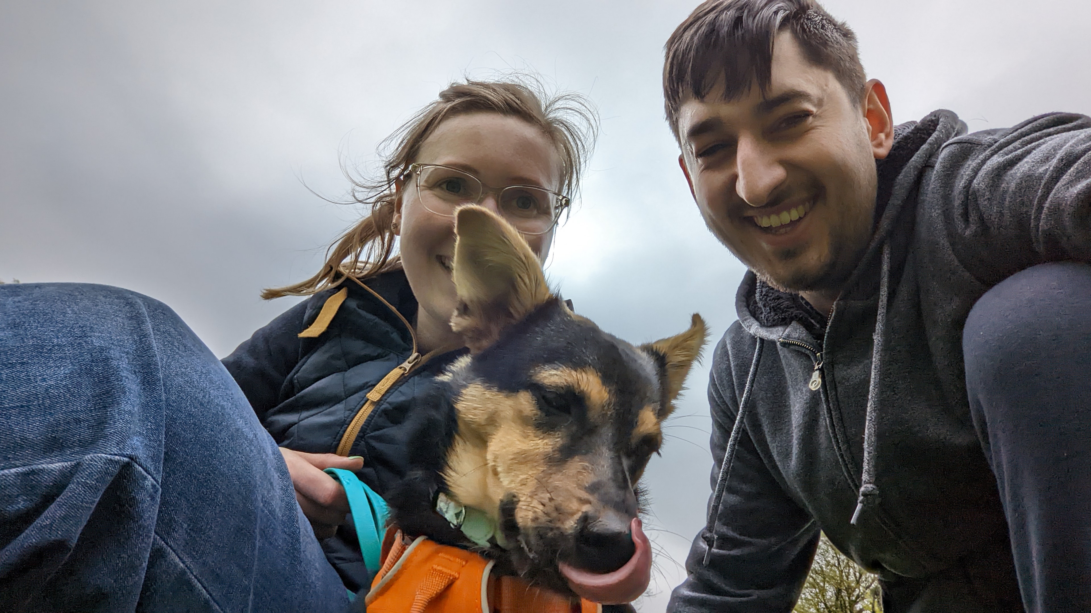 Selfie of Anna, Cookie and Jamie. Anna's face is partially obscured by Cookie the dog, who's in the middle of licking her lips, and looking slightly away from camera. Anna and Jamie are both smiling at the camera
