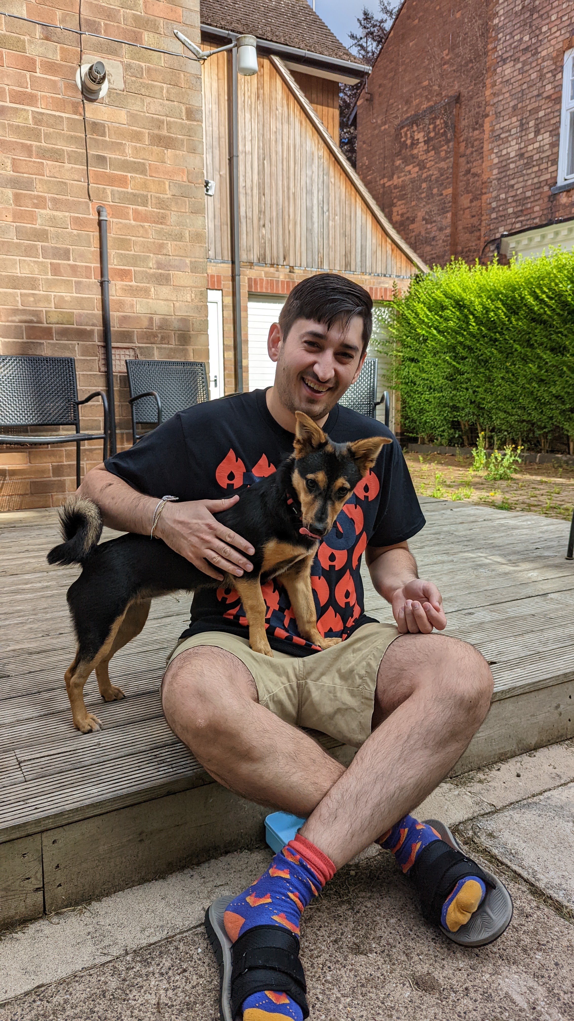 Jamie and Cookie sitting outside on the decking, with Cookie half on Jamie's lap, with her tongue in a mlem as she's just eaten a biscuit to bribe her into shot. Jamie is smiling at the camera and wearing an incident.io t-shirt with lots of fires (their logo)