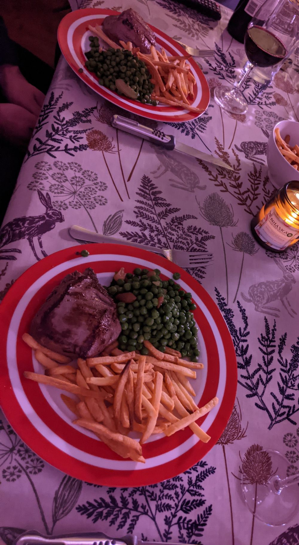 A slanted photo of a table set with two plates with a fillet steak (cooked blue / very very rare), minted peas, french fries, a bowl of overflow fries, and a large glass of Malbec