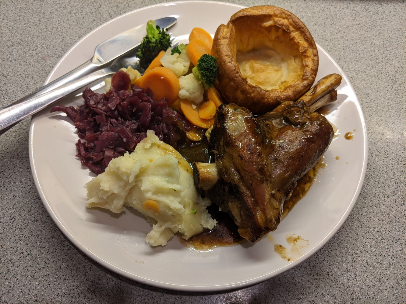 A plate with a lamb shank, with cheddar mashed potatoes, red cabbage, broccoli, cauliflower, carrots, a Yorkshire pudding, and mint gravy