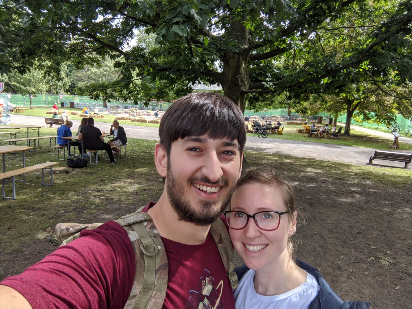 Selfie of Anna and Jamie, standing outside in the sunny Arboretum, with people socially distanced at tables and deckchairs