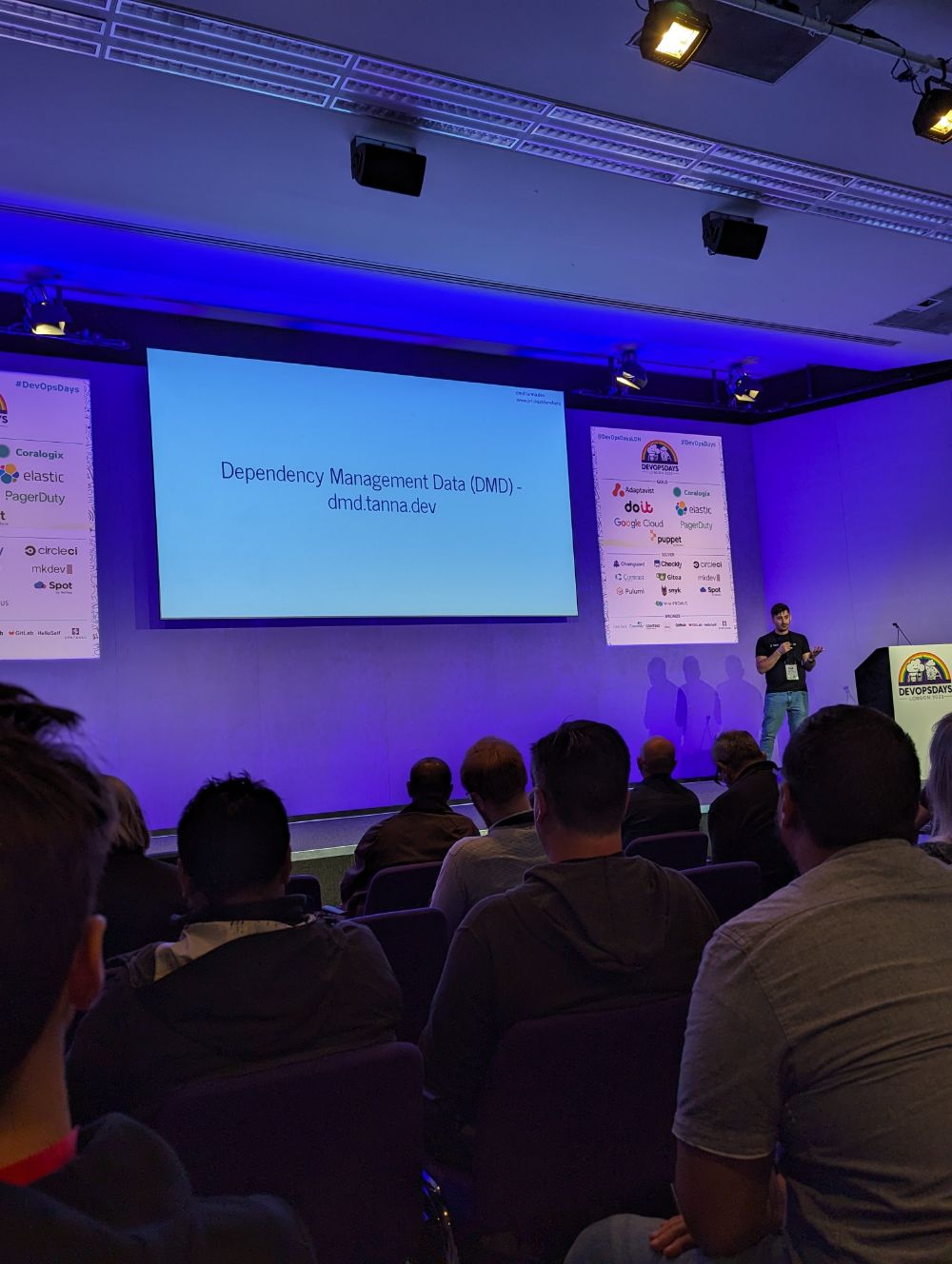 Jamie speaking on stage at DevOpsDays London, in front of a slide about the creation of the Open Source project dependency-management-data (dmd.tanna.dev)