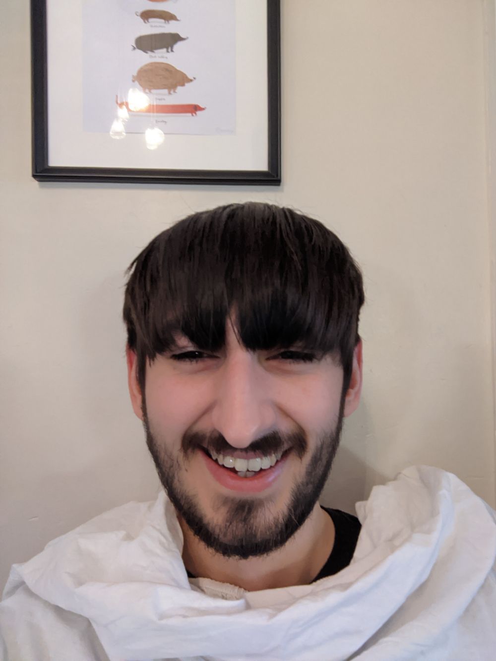 Selfie of Jamie before the haircut, with a white sheet around him to catch the falling hair, with a lot of beard and moustache growth, and hair slightly covering his eyes