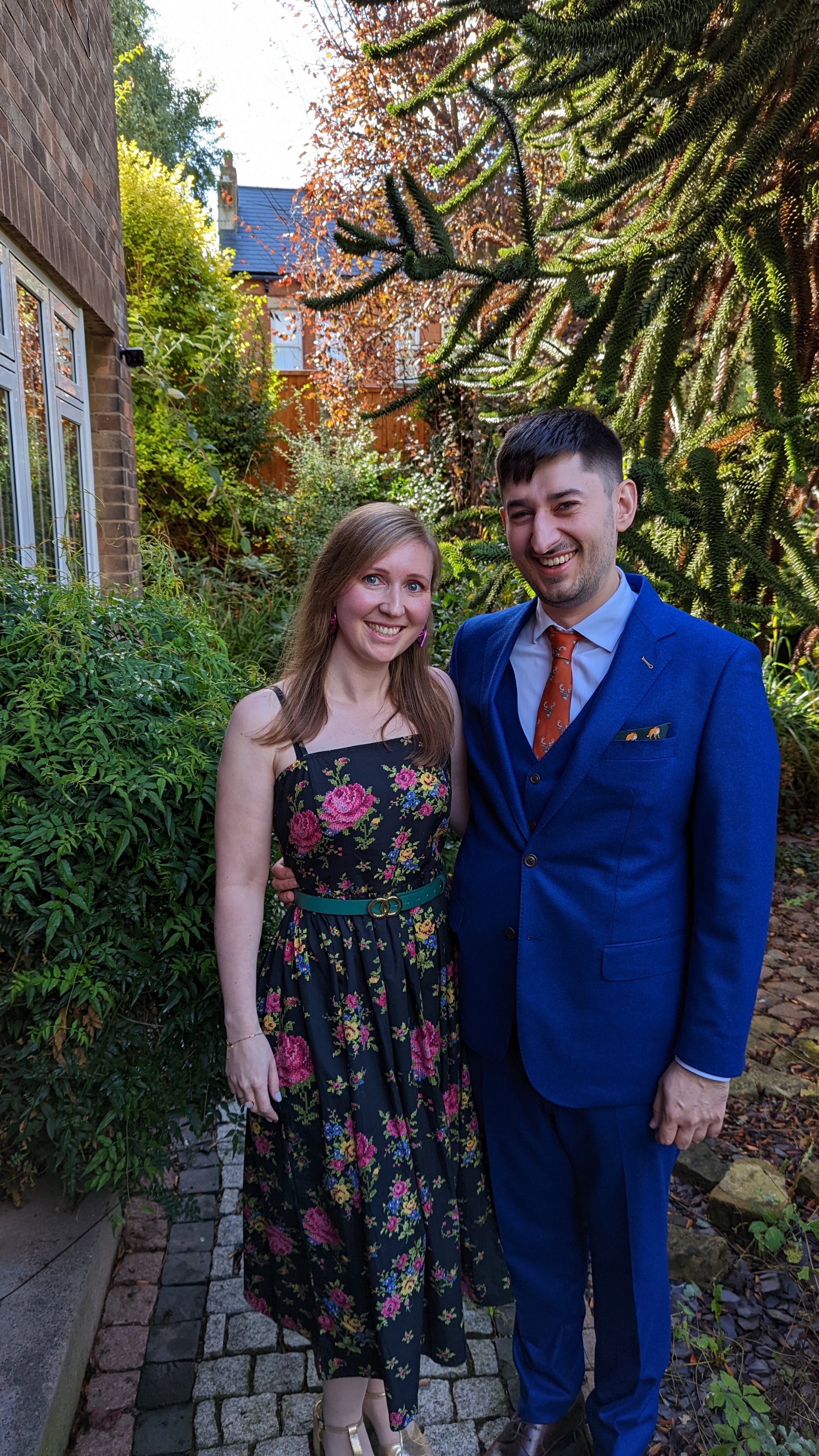 Anna and Jamie standing outside their house, ahead of their friends' wedding, looking very cute.

Anna is looking beautiful  in a flowery dress, with gold shoes. She has a pair of purple heart earrings 

Jamie is in a custom made blue suit, from Black Butterfly in Nottingham, with brown shoes and mustard socks (not pictured), a bronze tie with stags on it, and a green pocket square with foxes on it.

Behind them you can see their monkey puzzle tree.
