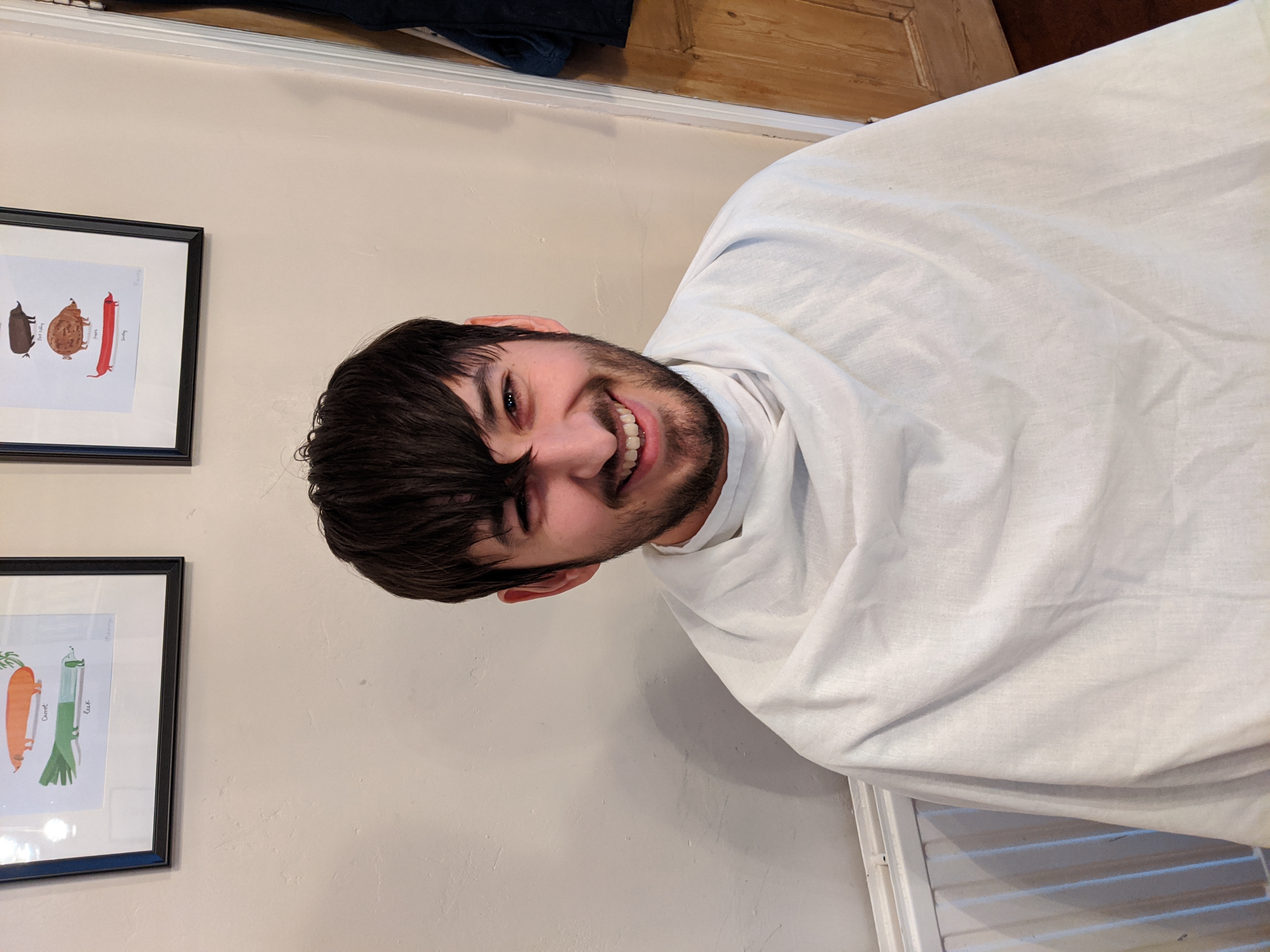 Photo of Jamie sitting on a chair with a bed sheet around him, smiling a little apprehensively, with long hair in his eyes